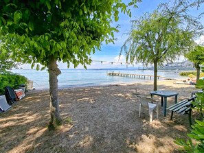 A beach with a wooden pier extending into the water, a table and chairs under a tree, and a grill set up on the sand.