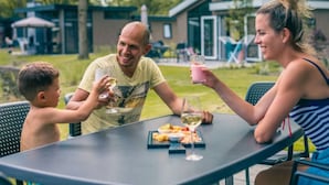 A family dining outdoors with a table set for a meal, chairs, and a building in the background. - Panorama Cottage 4 (Kaatsheuvel)