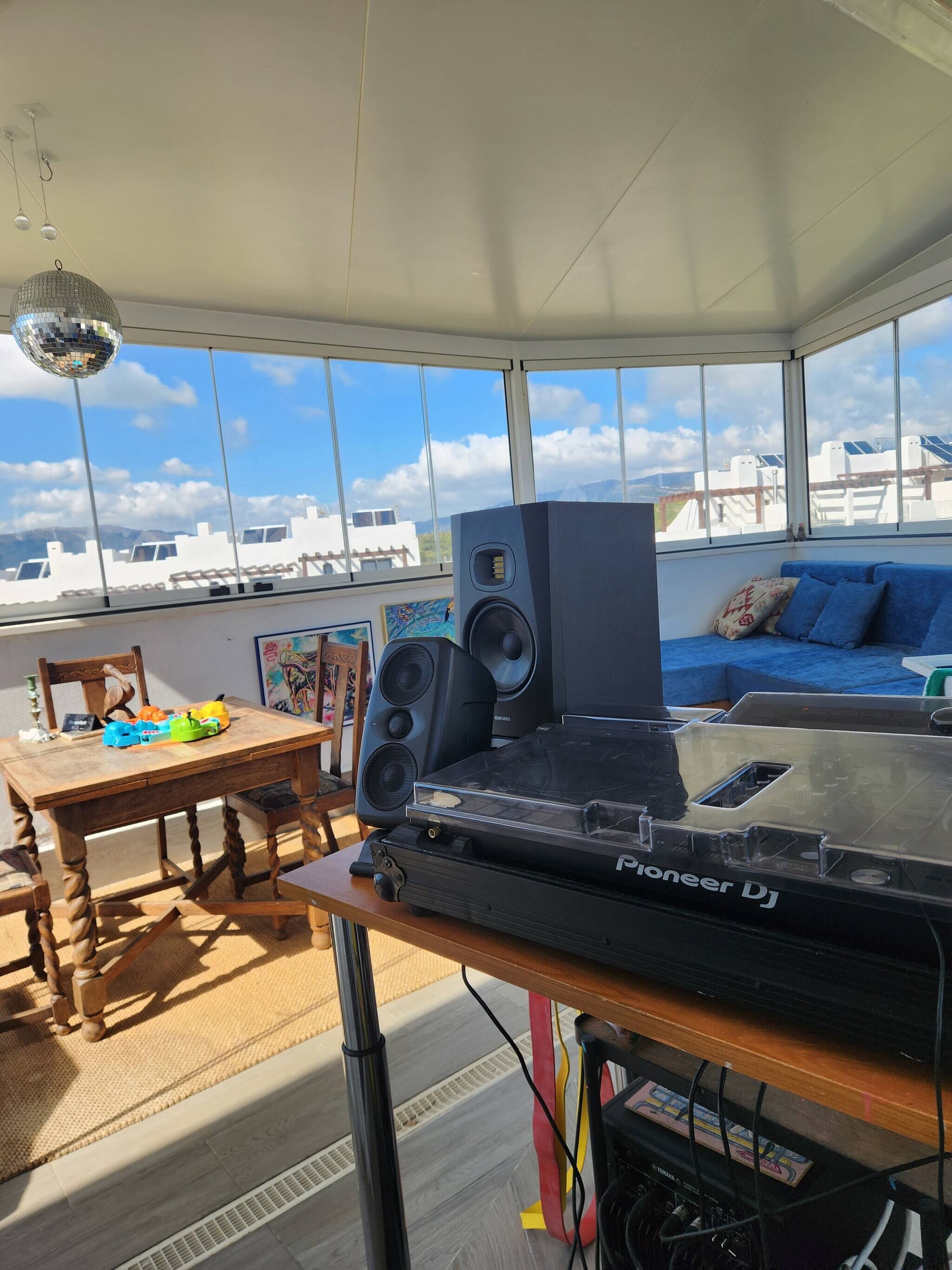 Um toca-discos Pioneer numa mesa de madeira em um quarto iluminado pelo sol, com janelas grandes, um sofá azul e uma bola de espelhos.
