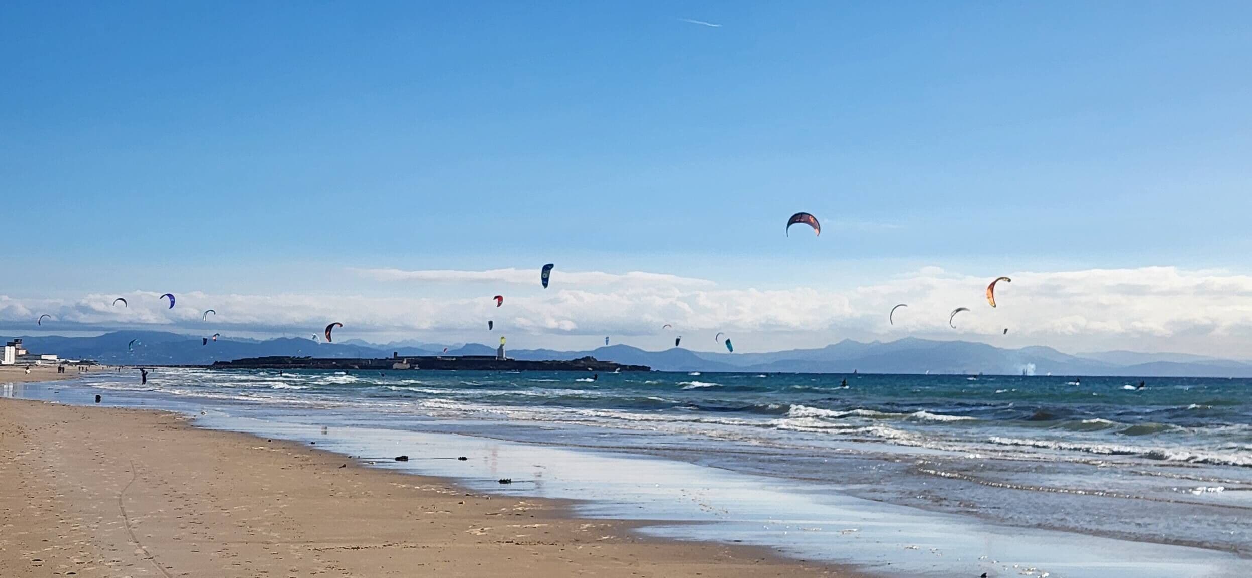 Uma praia com pipas voando, céu claro e montanhas ao fundo.