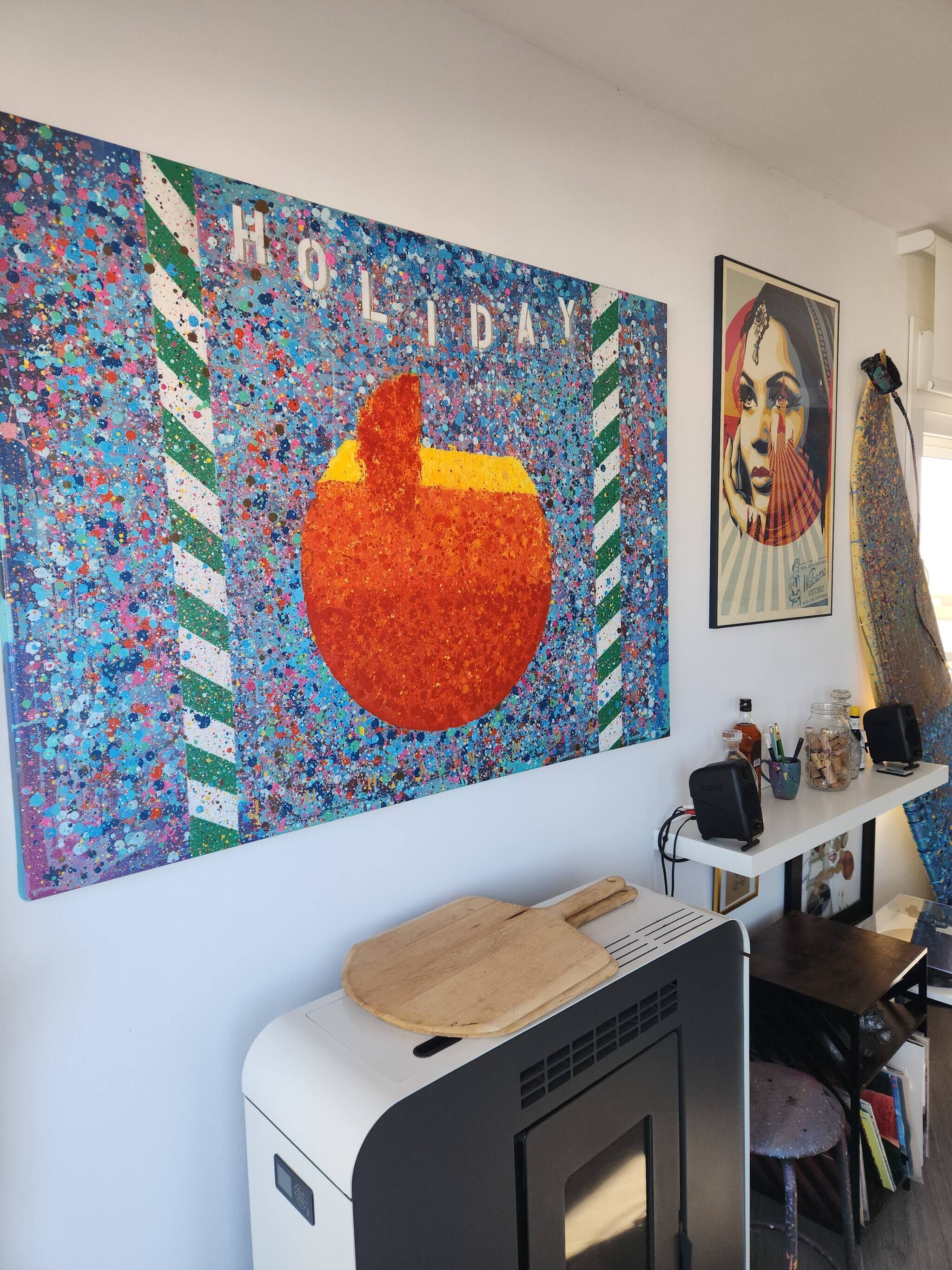 Quarto com uma pintura abstrata colorida, um retrato emoldurado e uma escrivaninha com computador e colunas de som.