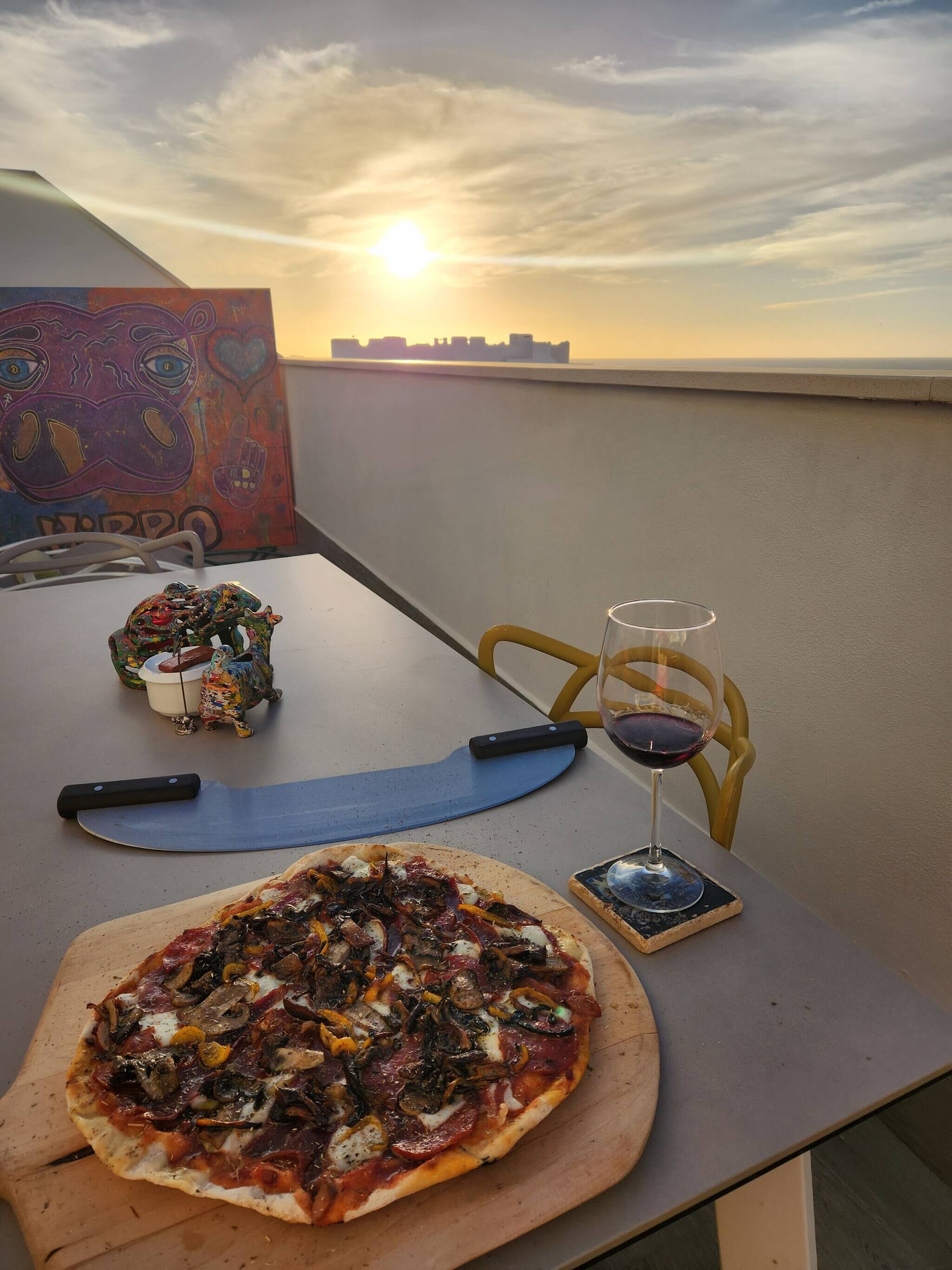 Pizza em tábua de madeira, cortador de pizza, taça de vinho com vinho tinto, e uma cadeira em mesa no terraço com vista para o pôr do sol.