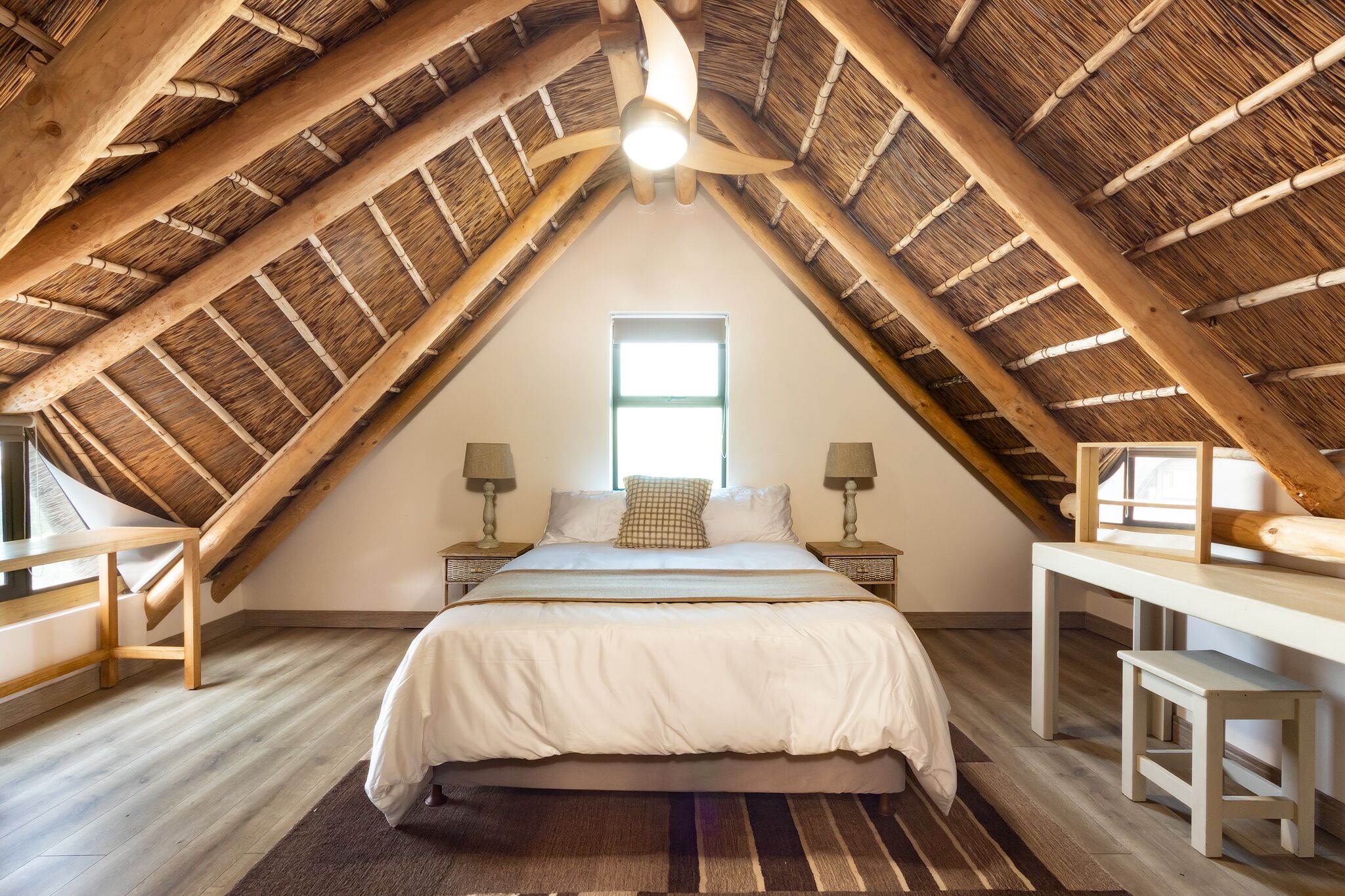 Quarto com teto inclinado, uma cama, mesas de cabeceira, uma janela e uma escrivaninha.