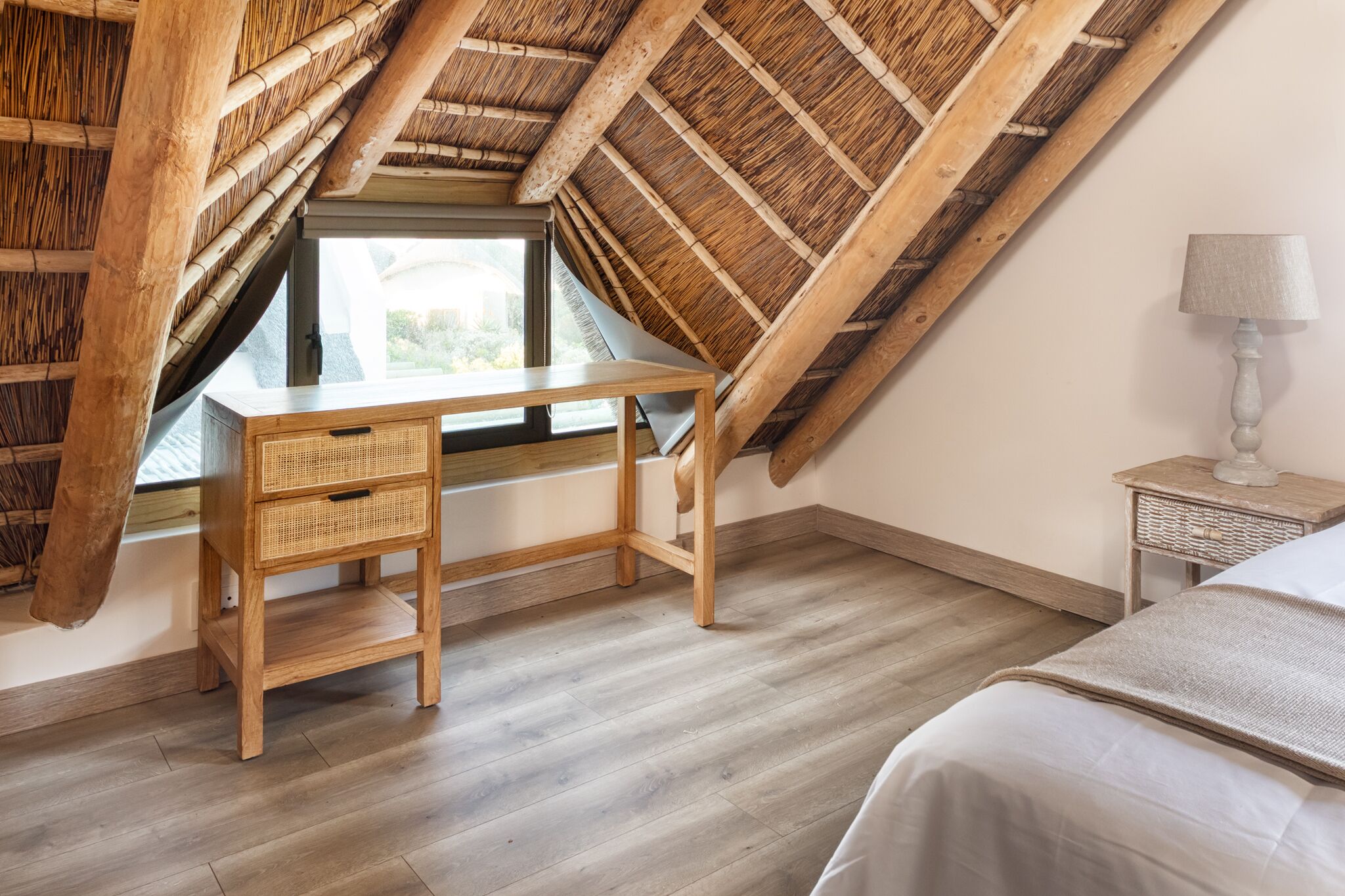 A room with a sloped ceiling, a wooden desk with drawers, a bedside table, and a lamp.