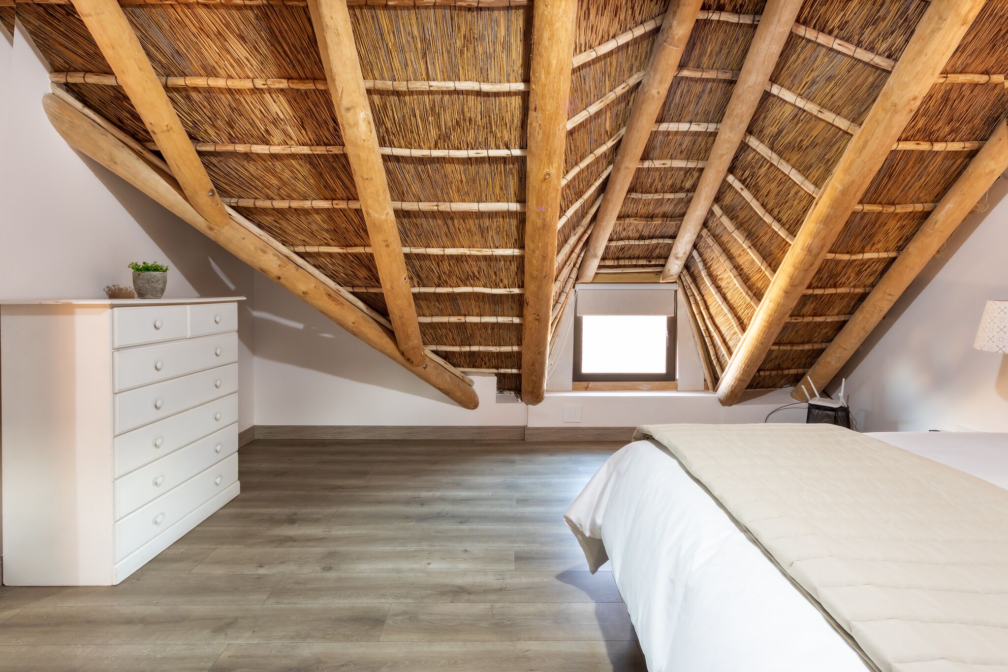 A room with a sloped ceiling, wooden beams, a bed, a dresser, and a small window.