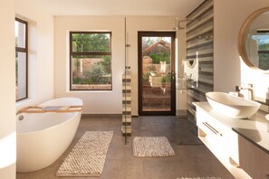 Ein modernes Badezimmer mit einer freistehenden Badewanne, einem großen Spiegel und Blick ins Freie durch eine Glastür.