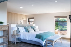 Un dormitorio moderno con una cama grande, mesitas de noche, un banco y vistas a la vegetación por la ventana.