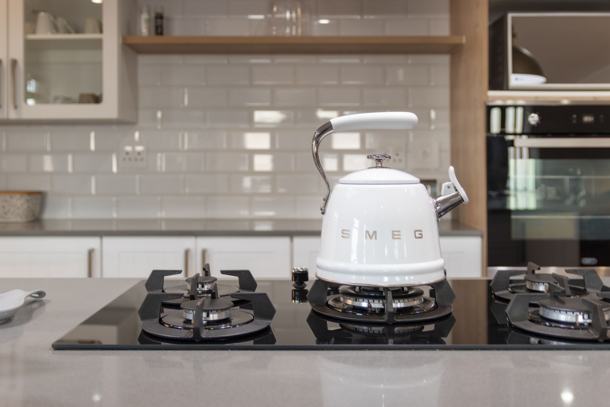 A kitchen with a white electric kettle on a modern stove, white tiled backsplash, and stainless steel appliances.