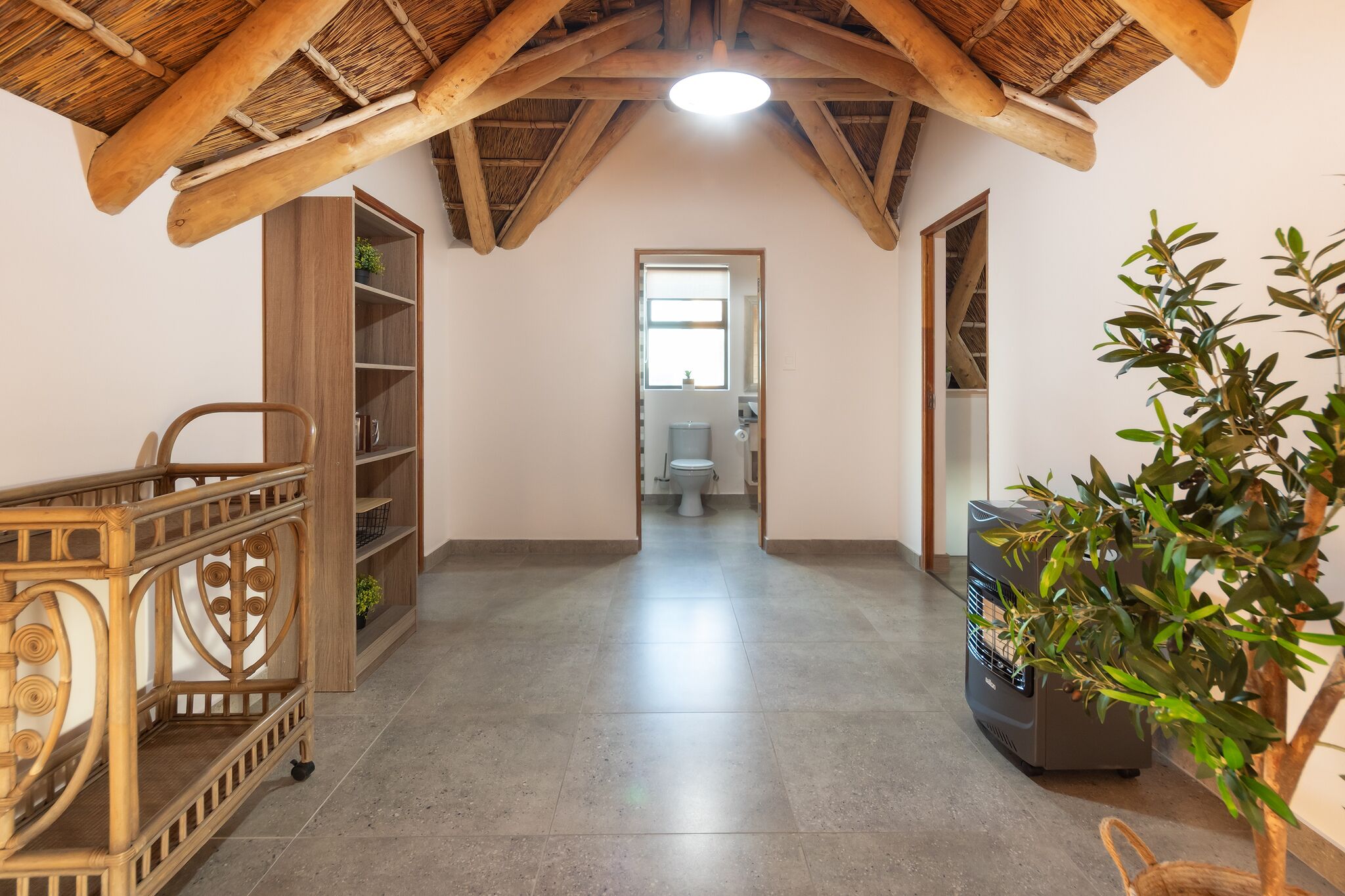 A room with a high ceiling, wooden beams, a central door leading to a bathroom, a wicker chair, and a potted plant.