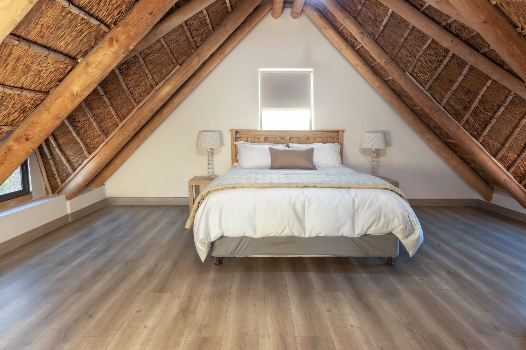 Een slaapkamer met een schuine kap, een bed met houten hoofdbord, bedlampen en een raam.