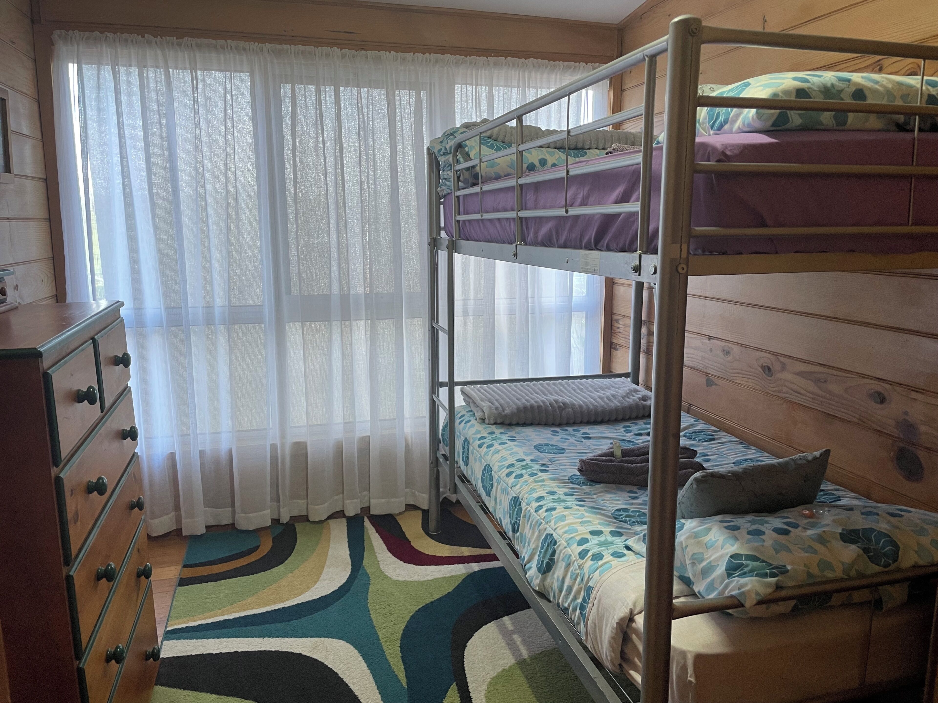 A bunk bed room with a wooden dresser, a colorful rug, and sheer curtains.