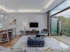 Ein modernes Wohnzimmer mit Flachbildfernseher, einer Couch, einem Essbereich mit Stühlen und einem großen Fenster mit Blick ins Freie.