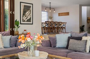 Ein Wohnzimmer mit einer Couch, einem Couchtisch mit Blumenvase, einem Essbereich mit Stühlen, einer Wandleuchte und einem gerahmten Bild.