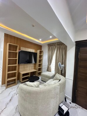 Una sala de estar moderna con un televisor de pantalla plana, estanterías empotradas de madera, un sofá blanco, una mesita pequeña y un ventilador blanco.
