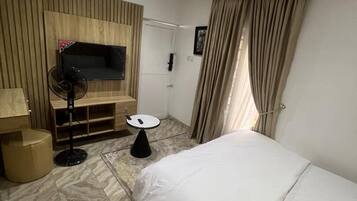 A hotel room with a bed, a TV, a fan, a small table, and an air conditioning unit.