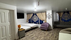 A bedroom with a bed, a chair, a sofa, a TV, and a door. - Standard Double Room (Bolton)