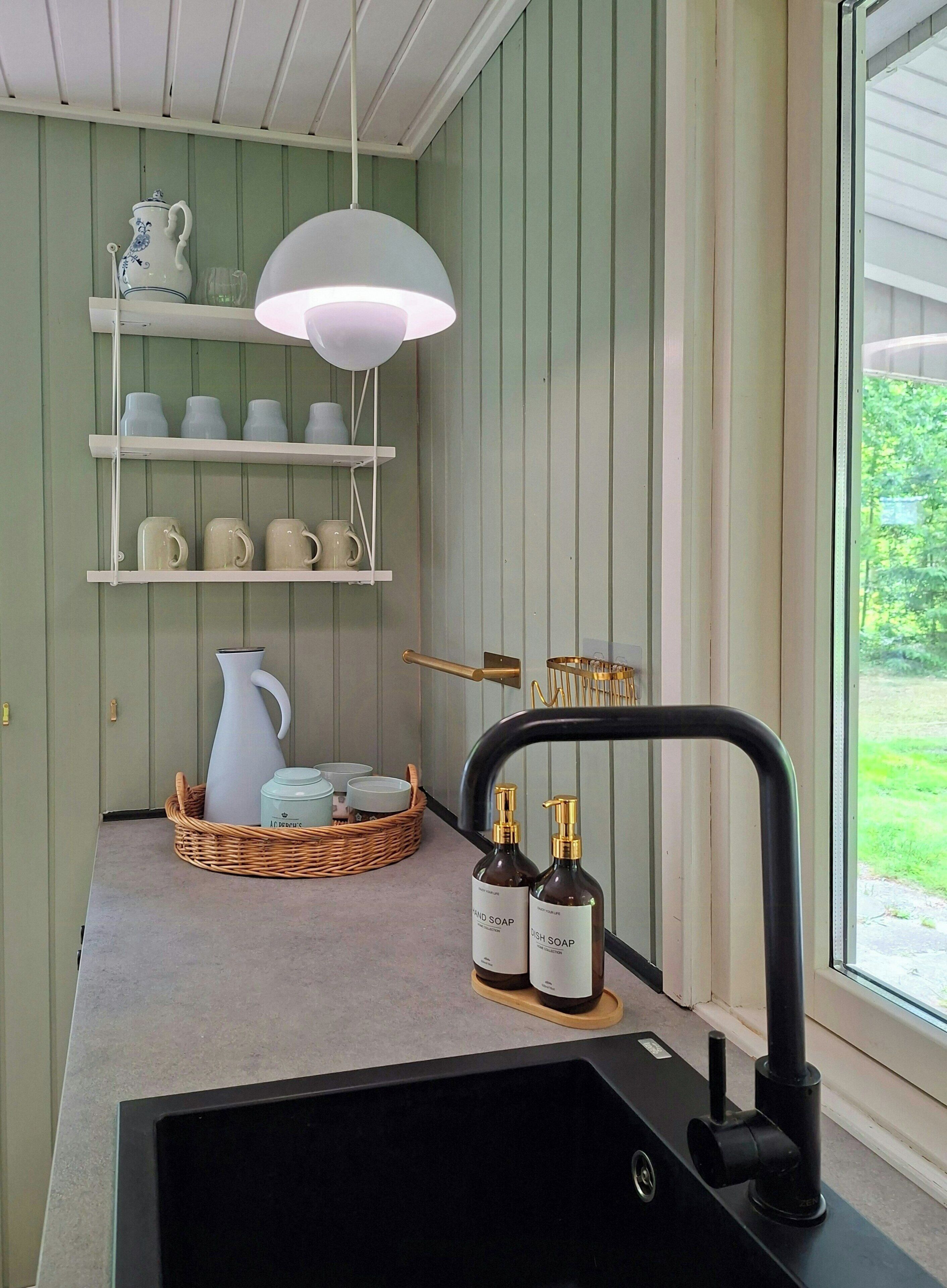 A kitchen with a black sink, a white pendant light, shelves with mugs, and a window with a view of greenery.