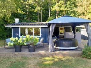 Eine Holzterrasse mit überdachter Sitzgruppe, einem kleinen Gartenhaus und Kübelpflanzen.