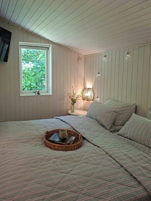 Ein Schlafzimmer mit einem großen Bett, einem Nachttisch mit Lampe, einem Fenster mit Blick ins Grüne und einem an der Wand befestigten Fernseher.
