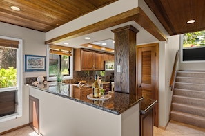 A kitchen with granite countertops, wooden cabinets, and a central island with a wine bottle and grapes. - Two-Story Ocean View Retreat Steps from Pool & Beaches -KRV 915 (Lahaina)