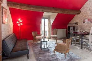 Ein gemütlicher Raum mit roter Decke, einer Couch, zwei Korbstühlen, einem kleinen runden Tisch und einem Esstisch mit Stühlen.