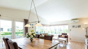 Amplia habitación con una mesa de comedor de madera, dos sillas, un sofá y una mesa de centro. Grandes ventanales ofrecen luz natural y vistas al verdor exterior.