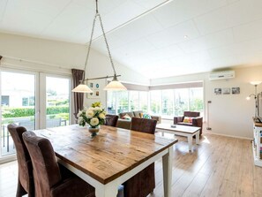 A spacious room with a wooden dining table, two chairs, a sofa, and a coffee table. There are large windows providing natural light and a view of greenery outside. - Holiday Home in Noordwijk With Terrace (Noordwijk)