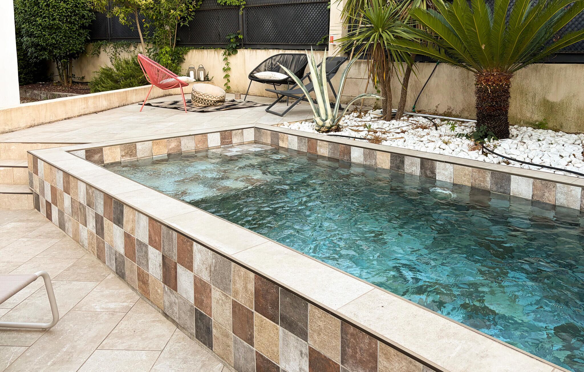 A rectangular pool with tiled edges, surrounded by a patio area with seating and plants.