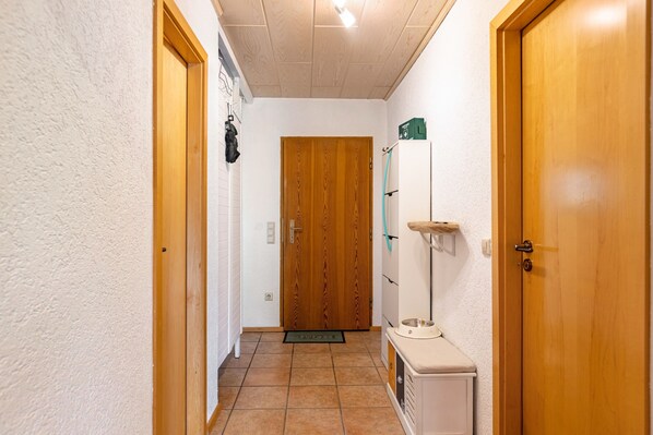 A narrow hallway with a wooden door at the end, a white wall, and a tiled floor.
