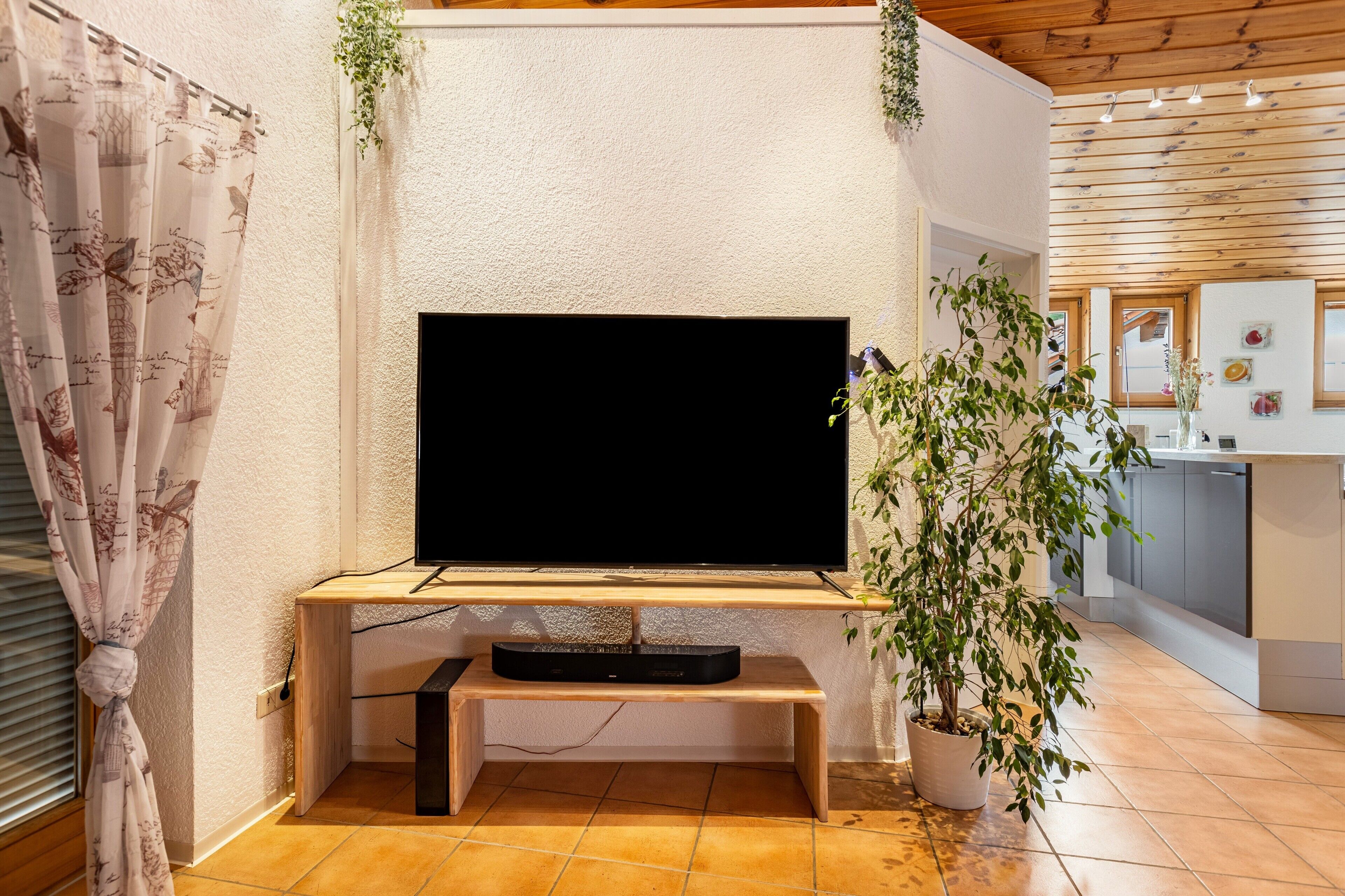 Ein Wohnzimmer mit einem an einer Holzständerung befestigten Flachbildfernseher, einer Topfpflanze und einem Fenster mit Vorhängen.