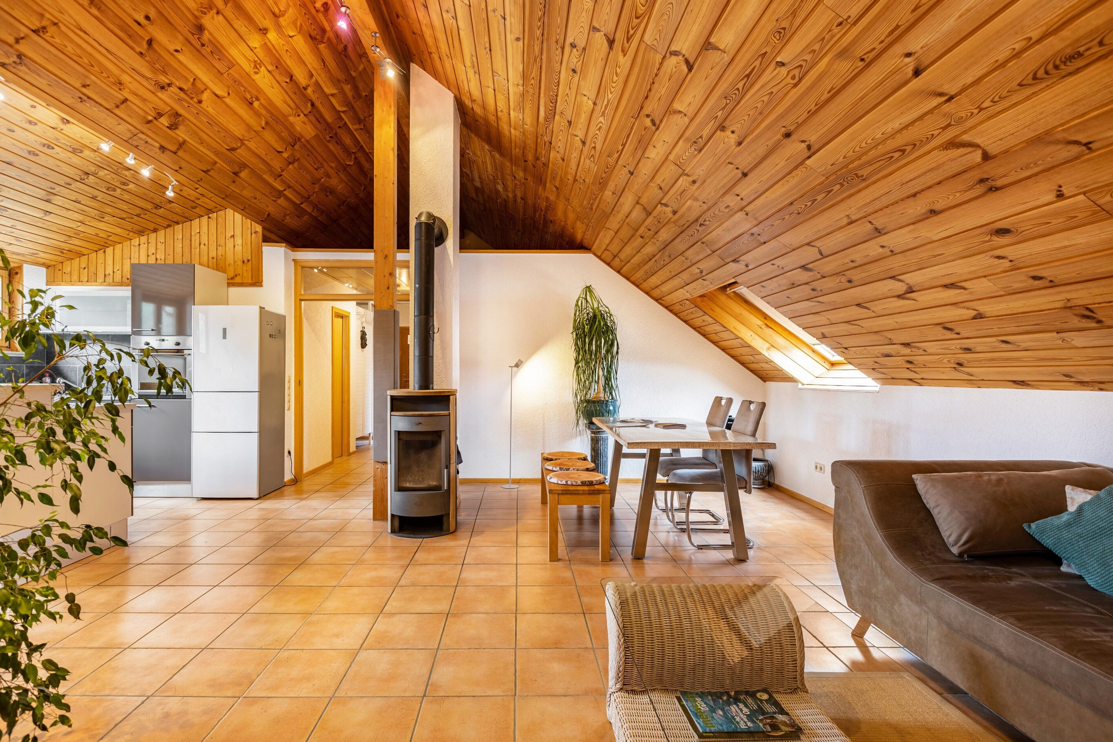 Ein großzügiger Wohnbereich mit hoher Holzbalkendecke, einem zentralen Kamin, einem Esstisch mit Stühlen, einer Couch und einer Küchenzeile mit Geräten.