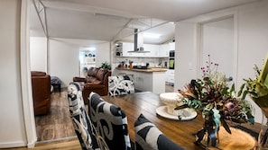 Een moderne keuken met witte kasten, een eethoek met een houten tafel en gestreept beklede stoelen, en een woonkamer met een bruine leren bank en een vloerkleed met dessin.