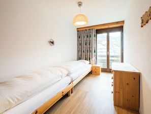 Ein Einzelbett mit Holzbettgestell, ein kleiner Nachttisch, ein Schreibtisch mit Schubladen, eine Pendelleuchte und ein Fenster mit Vorhängen.