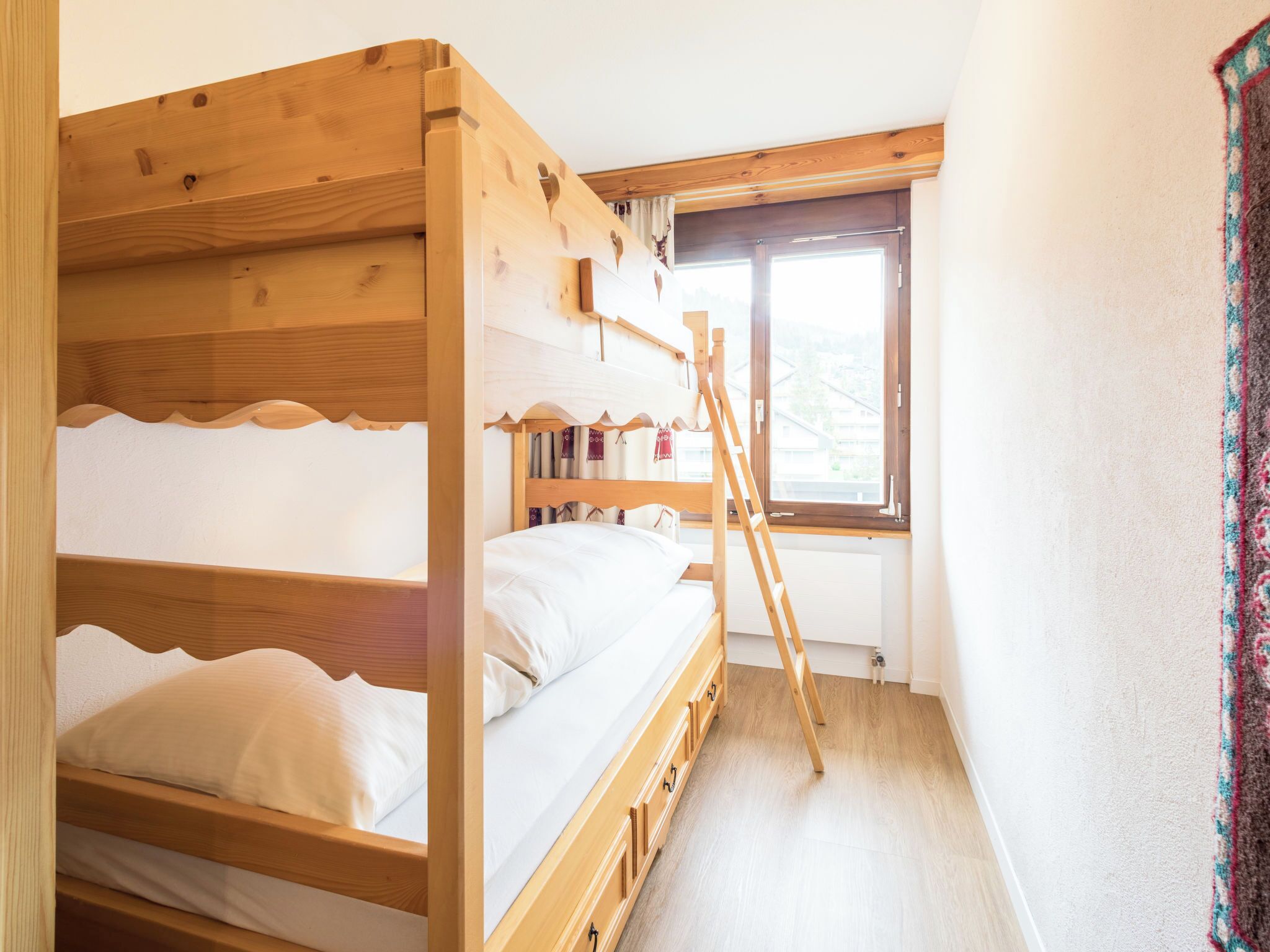 A wooden bunk bed room with a ladder, a window with curtains, and a view of buildings outside.