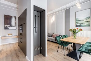 A modern kitchen with a built-in oven, a dining area with a wooden table and green chairs, and a gray sofa in the background. - Brand-new, designer 3-bedroom apartment (Berlin)