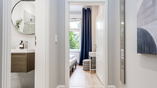Un pasillo que conduce a un dormitorio con ventana, espejo y un baño con lavamanos y espejo.