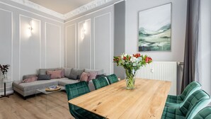 Una sala moderna con una mesa de comedor de madera, sillas tapizadas en verde, un sofá gris con cojines, un jarrón de flores y un cuadro en la pared.