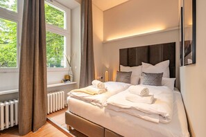 Ein ordentlich bezogenes Bett mit weißen Leinen und Handtüchern, ein dunkles Kopfteil sowie ein Fenster mit Vorhängen.
