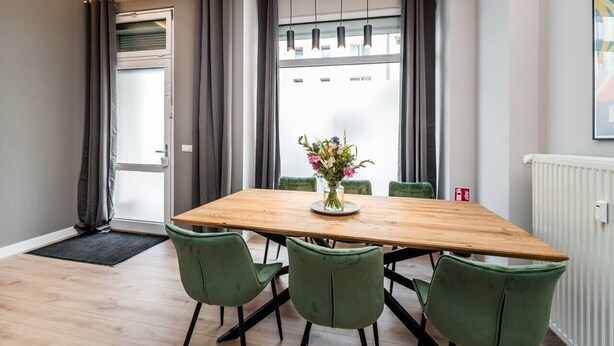 A dining area with a wooden table, green chairs, a vase with flowers, and a view of buildings through large windows. - Luxurious 3-Bedroom Designer Apartment (Berlin)