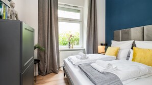 A modern hotel room with a bed, a nightstand, a window with curtains, a radiator, and a bookshelf with books. - Luxurious 3-Bedroom Designer Apartment (Berlin)