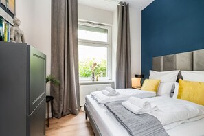 A modern hotel room with a bed, a nightstand, a window with curtains, a radiator, and a bookshelf with books. - Luxurious 3-Bedroom Designer Apartment (Berlin)