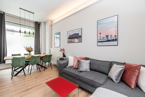 A modern living room with a grey sofa, green chairs, a red ottoman, and framed cityscape pictures on the wall. - Luxurious 3-Bedroom Designer Apartment (Berlin)