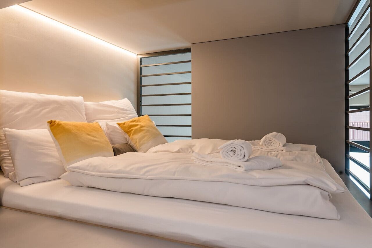 A modern hotel room with a neatly made bed, a window with blinds, and a wall-mounted light fixture.