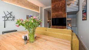 Un salon moderne avec une table à manger en bois, un canapé jaune, un grand téléviseur et un mur en briques.