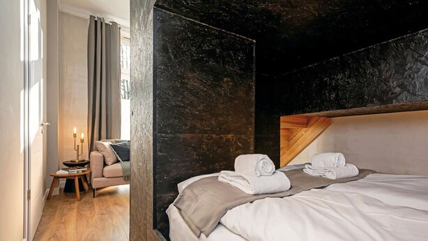 Una habitación de hotel moderna con una cama grande, un cabecero de madera, una mesita de noche con una lámpara y vistas a un paisaje urbano a través de una ventana.