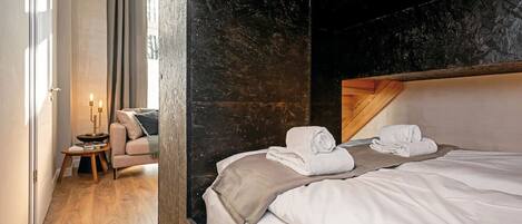 A modern hotel room with a large bed, a wooden headboard, a small side table with a lamp, and a view of a cityscape through a window.