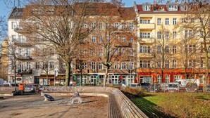 Una hilera de edificios de varios pisos con balcones, una zona pavimentada con un banco circular y coches aparcados.