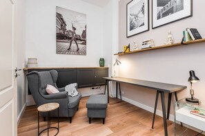 Ein modernes Wohnzimmer mit einem grauen Sofa, einem kleinen runden Tisch, einem Glastisch und einem Schreibtisch mit Lampe. An den Wänden hängen gerahmte Bilder.
