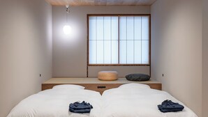 Una habitación con techo de madera, un ventanal con paneles shoji, dos camas con ropa blanca y un reposapiés redondo pequeño.