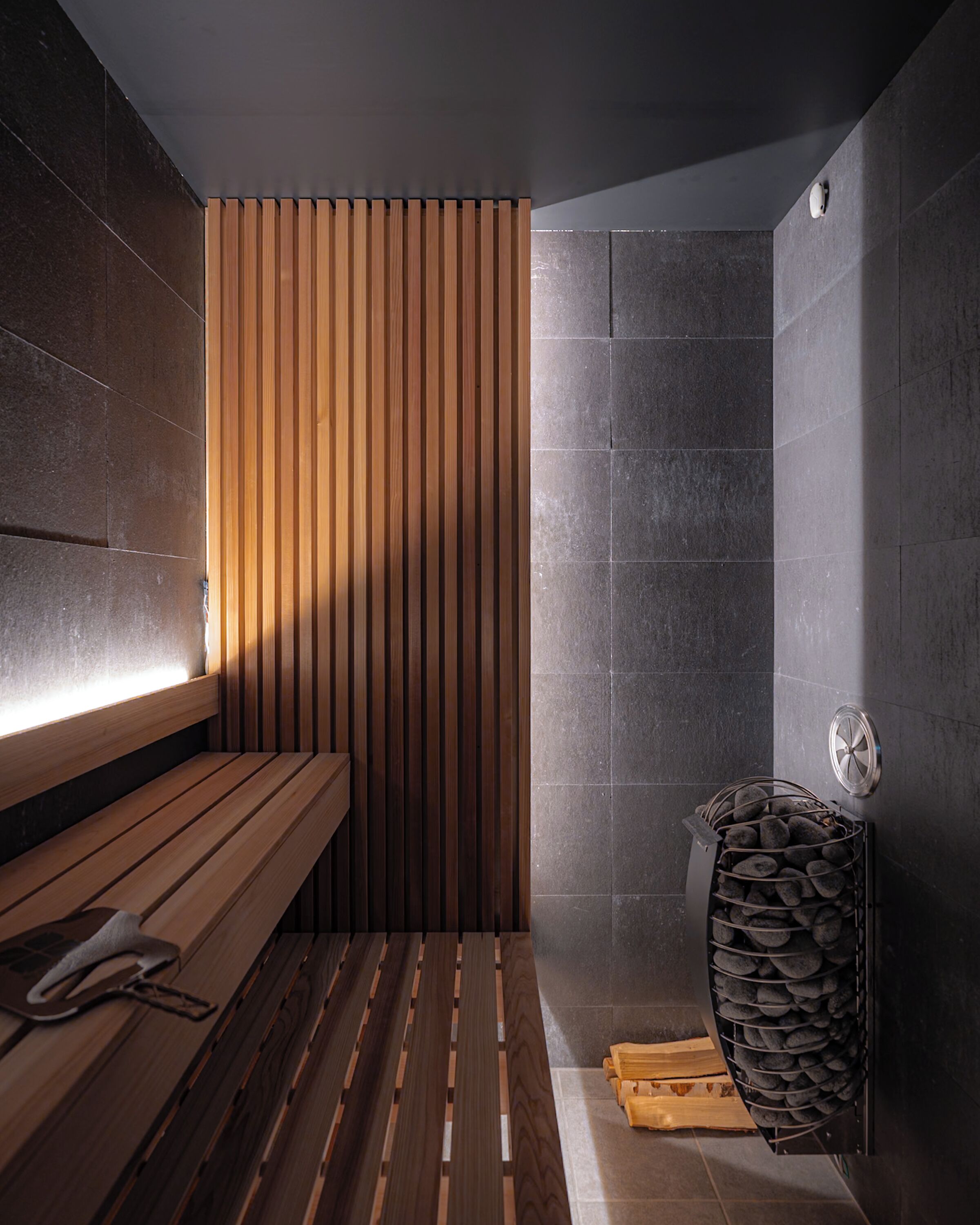 A modern sauna with wooden benches, a stone wall, and a metal rack for towels.